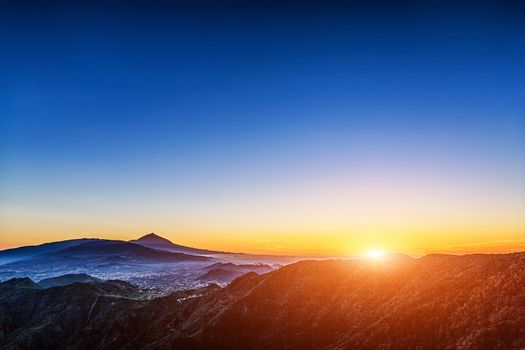 Sun with sunlight over mountains on blue sky with fog and Teide volcano on background at evening sunset in Tenerife Canary island, Spain