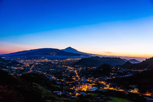 City or town with illumination after sunset or sundown at evening in mountains with blue sky and Teide volcano on background in Tenerife Canary island, Spain at spring or summer