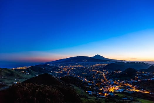 City or town with illumination after sunset at evening in mountains with blue sky and Teide volcano on background in Tenerife Canary island, Spain