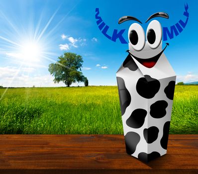 White carton milk with black spots with text Milk in the shape of horns. In a countryside landscape with green grass and a tree on blue sky with clouds and sun rays