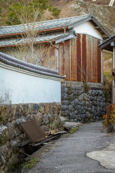 A rusted garage near a curving path and some trash against a white wall with traditional Japanese roof tiles.