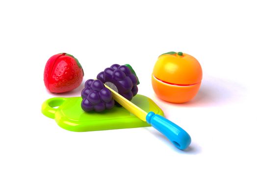 A plastic toy strawberry, mandarin orange and grapes on a cutting board being cut in half with a plastic knife.