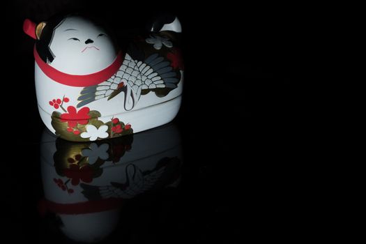A Japanese Ceramic cat decorated with flowers and a crane reflected in glass on a pure black background.