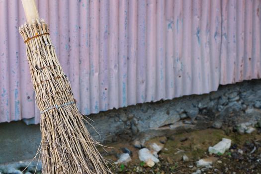 A straw bamboo broom leaning against an old wall outside.