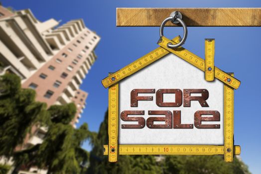 Yellow wooden meter ruler in the shape of house with text for sale. For sale real estate sign with tall and blurred buildings in the background
