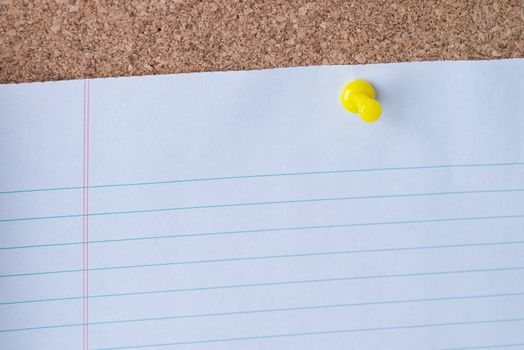 A yellow thumb tack holding a white piece of lined loose leaf paper on a cork board.
