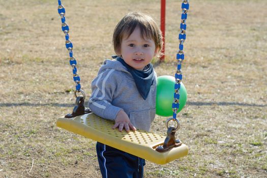 A 2 year old boy holding a ball and standing by the swings at a playground.