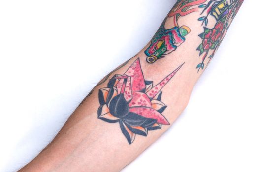 A tattoo of an origami pink crane with a cherry blossom pattern on a black lotus surrounded by bits of other tattoos on a forearm.