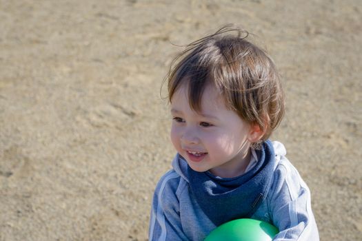 A 2 year old boy holding a green ball smiling in a playground.