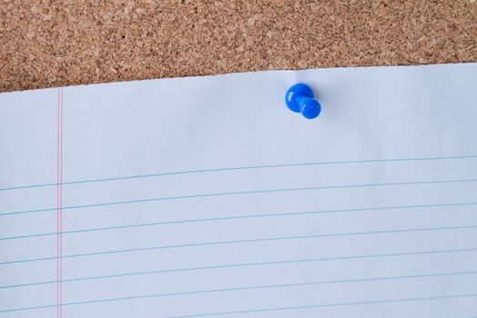 A blue thumb tack holding a white piece of lined loose leaf paper on a cork board.