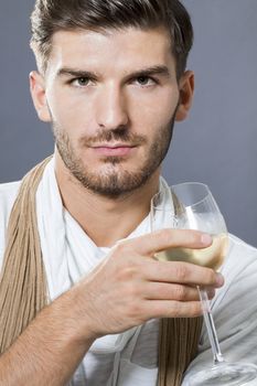 Sexy handsome young man with a beard wearing an elegant scarf drinking white wine and looking at the camera with a serious intense expression