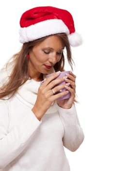 Cold attractive young woman with a cute smile in a festive red Santa hat sipping a hot mug of coffee that she is cradling in her hands to warm up in the winter weather, on white