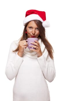 Cold attractive young woman with a cute smile in a festive red Santa hat sipping a hot mug of coffee that she is cradling in her hands to warm up in the winter weather, on white