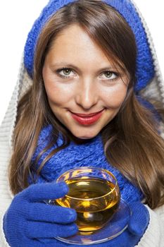 Fashionable young woman in a blue knitted winter ensemble and cowl neck jersey sipping a cup of hot tea with a smile in an effort to keep warm, isolated on white