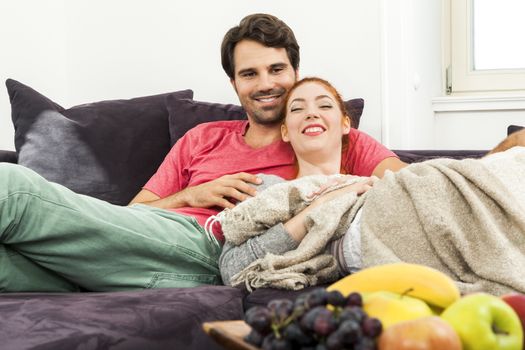 Young Couple Resting on the Sofa at the Living Room While the Man is Watching TV and Woman is Reading a Book.