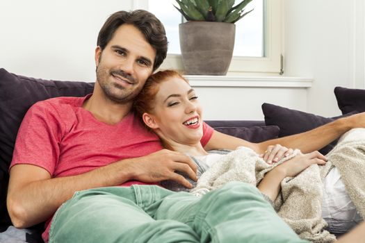 Young Couple Resting on the Sofa at the Living Room While the Man is Watching TV and Woman is Reading a Book.