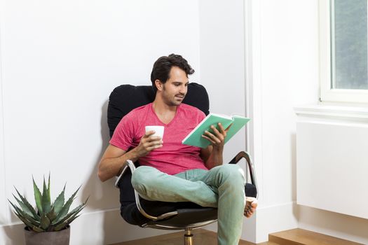 Young Man in Casual Clothing Sitting on Black Chair While Reading a Book and Holding a Glass of Drink.