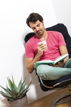 Young Man in Casual Clothing Sitting on Black Chair While Reading a Book and Holding a Glass of Drink.