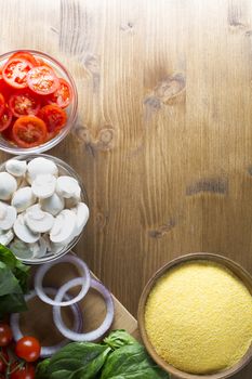 Ingredients for polenta recipe, with polenta in wooden bowl with sliced tomatoes, spinach and red onions.  Viewed from angle directly above with copy space on wooden table.  Can be used vertical or horizontal.