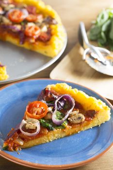 Vegan polenta pizza slice with cherry tomatoes, mushrooms, spinach and onion, on blue plate with whole pizza and ingredients out of focus in background giving room for copy.  Vertical orientation.