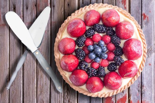 Fresh berries homemade tart with serving utensils, placed on a rustic wooden table. Above view.