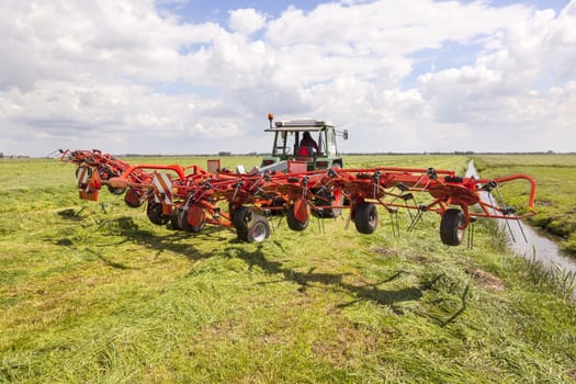 red hay turner behind tractor in green meadow in the netherlands