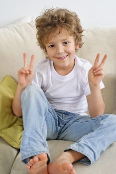 Young boy sitting on a sofa gives the V sign with both hands. His palms are towards the camera. He's looking at teh camera