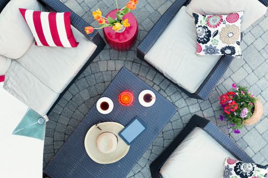 Relaxing on a brick patio by candlelight in the evening with two cups of tea and a straw sunhat on a table surrounded by deep seating comfortable armchairs and flowers, overhead view