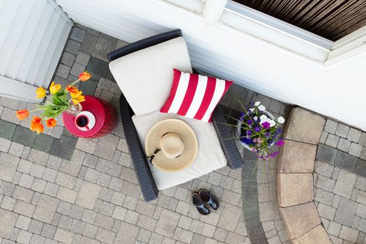 Taking a relaxing tea break in a deep seating patio set with a comfortable armchair flanked by colorful spring flowers with a sunhat and garden shoes on a brick paved outdoor patio, overhead view