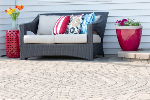 Comfortable modern deep seating settee on an outdoor brick paved patio with a circular pattern decorated with red ceramic flowerpot and pedestal table with colorful flowers