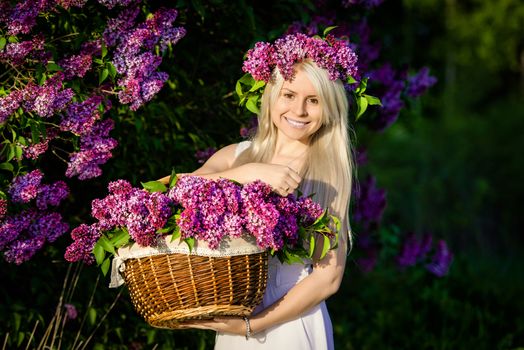 Beautiful smiling young woman is wearing wreath and basket of lilac flowers, white dress, sunset