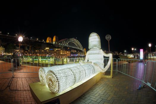 Dolly in foreground is a large 3-metre tall light-up doll made entirely from crochet and in the background, the SydneyHarbour Bridge and Park Hyatt  in background.  Tourists and people in motion.