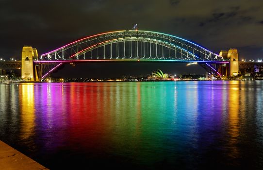 SYDNEY, AUSTRALIA - JUNE 3, 2013;  Sydney Harbour Bridge lit in vibrant rainbow colours and reflections in Sydney Harbour.  Sydney Opera House in far view.