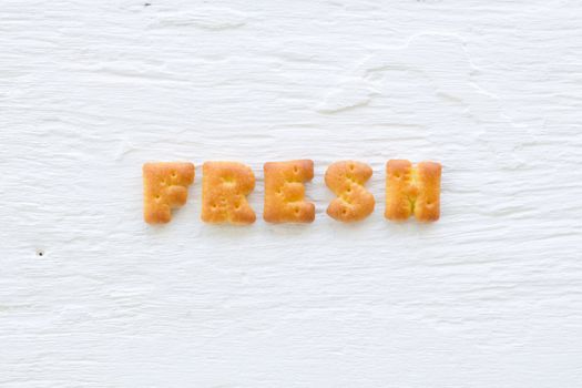 The upper-case letters word FRESH. Alphabet cookie crackers putting on rough texture of white wood background.