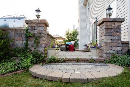 Welcoming entrance to an outdoor patio in front of a timber house with a decorative circular step from a green lawn between pillars with lamps to a patio with potted flowers and comfortable furniture