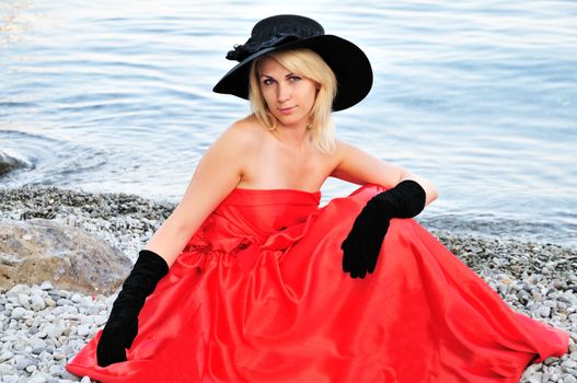 beautiful young woman wearing black hat, black gloves red dress is sitting on the beach near the sea 