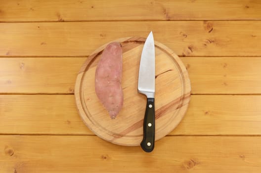 Whole raw sweet potato with a sharp kitchen knife on a wooden chopping board