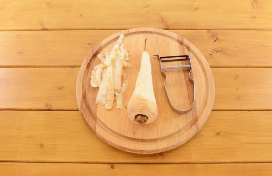 Raw parsnip being peeled with vegetable peeler on a wooden chopping board