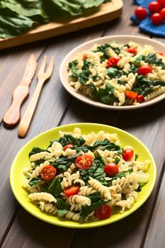 Two plates of fusilli pasta with chard leaves (lat. Beta vulgaris) and cherry tomatoes, photographed on dark wood with natural light (Selective Focus, Focus in the middle of the first dish)