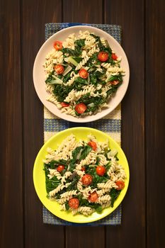 Overhead shot of two plates of fusilli pasta with chard leaves (lat. Beta vulgaris) and cherry tomatoes, photographed on dark wood with natural light