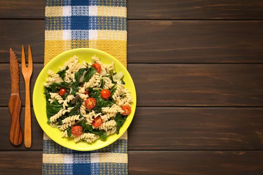 Overhead shot of fusilli pasta with chard leaves (lat. Beta vulgaris) and cherry tomatoes served on plate with wooden fork and knife on the side, photographed on dark wood with natural light