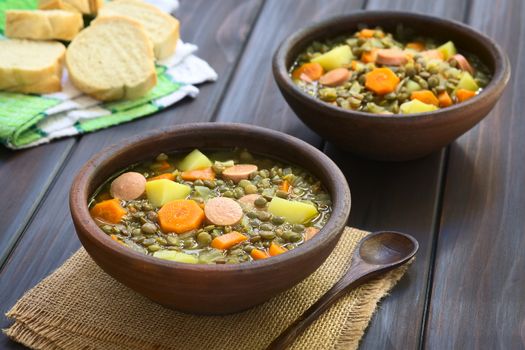 Two rustic bowls of lentil soup made with potato, carrot, onion and sausage slices, with slices of baguette bread in the back, photographed on dark wood with natural light (Selective Focus, Focus on the middle of the first soup)