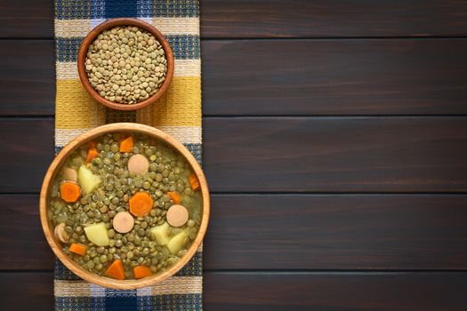 Overhead shot of a wooden bowl of lentil soup made with potato, carrot, onion and sausage slices, with small bowl of raw lentils above, photographed on kitchen towel on dark wood with natural light
