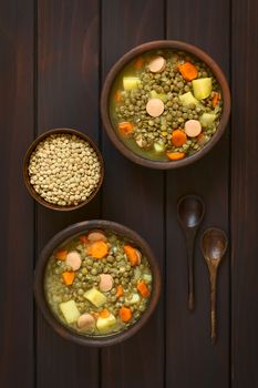 Overhead shot of two rustic bowls of lentil soup made with potato, carrot, onion and sausage slices, with a small bowl of raw lentils and wooden spoons on the side, photographed on dark wood with natural light