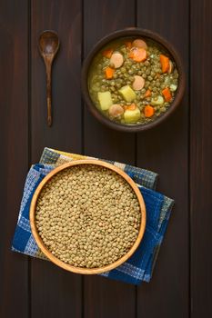 Overhead shot of a wooden bowl of raw lentils and a rustic bowl of lentil soup made with potato, carrot, onion and sausage slices, photographed on dark wood with natural light