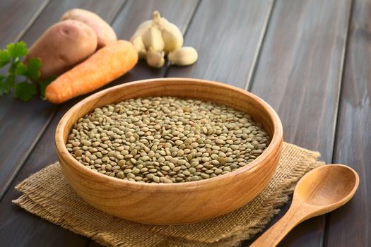 Raw lentils (lat. Lens culinaris) in wooden bowl with raw vegetables (potato, carrot, garlic) in the back, photographed on dark wood with natural light (Selective Focus, Focus one third into the raw lentils)