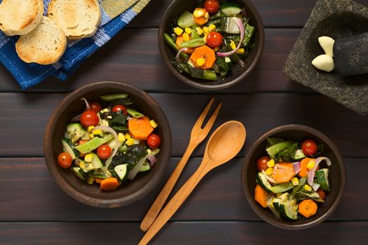 Overhead shot of three rustic bowls of baked vegetables (zucchini, onion, cherry tomato, broccoli, carrot, sweet corn, green bean, chard) seasoned with thyme, photographed on dark wood with natural light