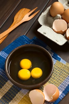 Three raw eggs in rustic bowl with egg box with eggs and eggshells in the back, photographed on dark wood with natural light (Selective Focus, Focus on the first two egg yolks)