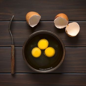 Overhead shot of three raw eggs in rustic bowl with fork on the side and broken eggshells above, photographed on dark wood with natural light