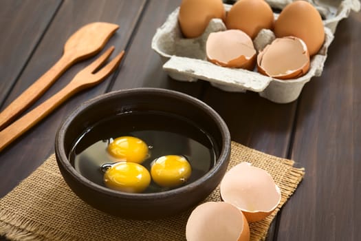 Three raw eggs in rustic bowl with egg box with eggs and eggshells in the back, photographed on dark wood with natural light (Selective Focus, Focus on the first two egg yolks)